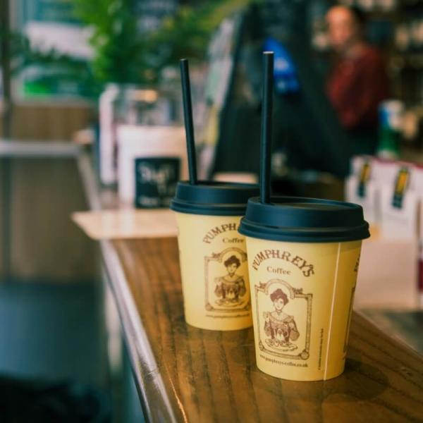 Two disposable Pumphrey's Coffee cups sit on top of a wooden counter. They are beige, with black plastic lids, and the Pumphrey's logo printed on their side. They have black paper straws sticking out of them.