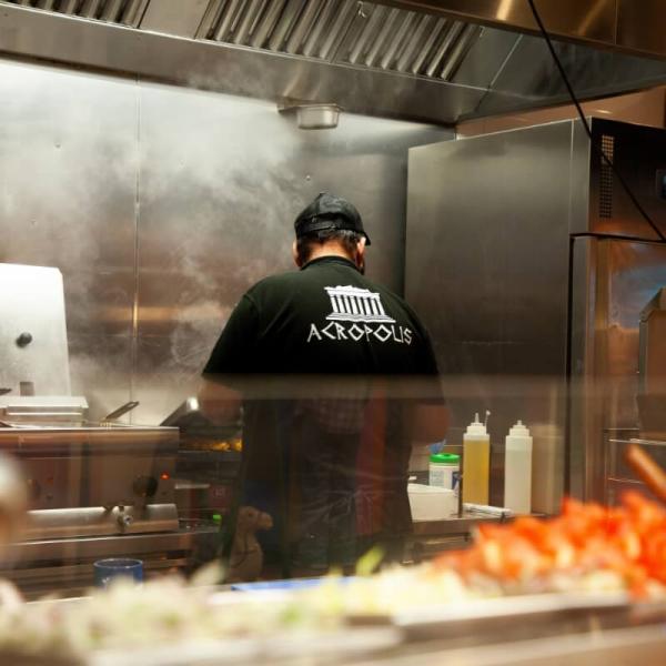 A man, wearing a green Acropolis t-shirt and a black baseball cap, faces away from the camera as he cooks food in Grainger Market